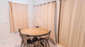 Una habitación con una mesa redonda de madera y cuatro sillas, cortinas color beige y piso de tonos claros.