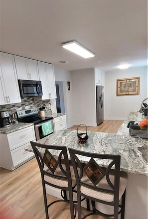 A modern kitchen with white cabinets, a microwave, stove, and a refrigerator. There is a dining area with a marble countertop and two chairs.