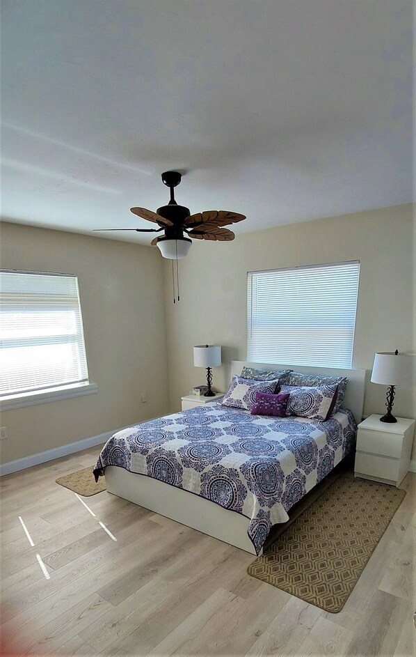 A bedroom with a bed, two bedside tables, a ceiling fan, and two windows with blinds.