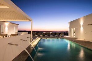 Una piscina en la azotea con sillones de descanso y vistas al horizonte de la ciudad al atardecer.