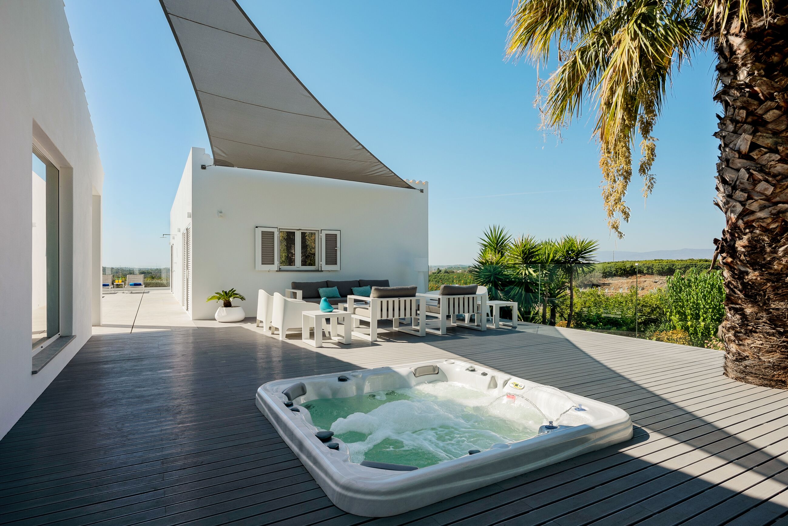 A modern outdoor deck with a hot tub, white furniture, and a view of the landscape.