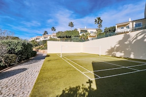 A tennis court with a net, surrounded by a paved walkway and lush greenery.