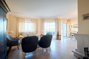 A spacious living room with a sofa, two armchairs, a dining table with chairs, and large windows with curtains.