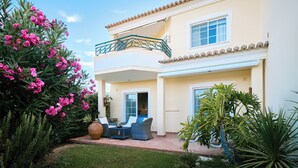Une maison à deux étages dotée d’un balcon, d’une terrasse avec des meubles d’extérieur et d’un jardin agrémenté de fleurs roses.