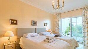 A hotel room with two beds, a chandelier, a window with curtains, and a painting on the wall.