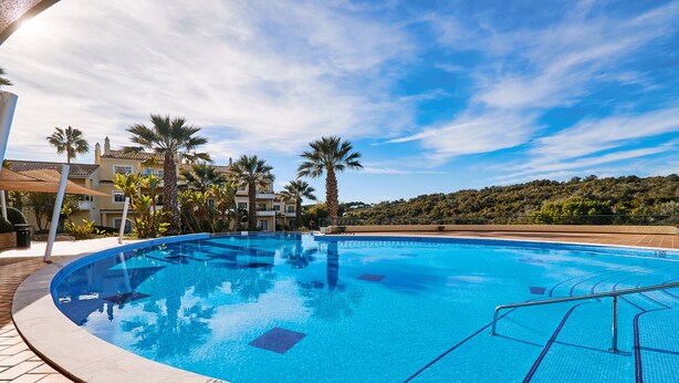 A clear blue swimming pool with tiled deck, surrounded by palm trees and a building with balconies.