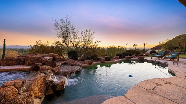 A pool with a waterfall feature, surrounded by rocks and desert vegetation, with lounge chairs and a view of the cityscape at sunset. - Camelback Heights - 8BR Estate w/ Pool, Theater & Camelback Views (Phoenix)
