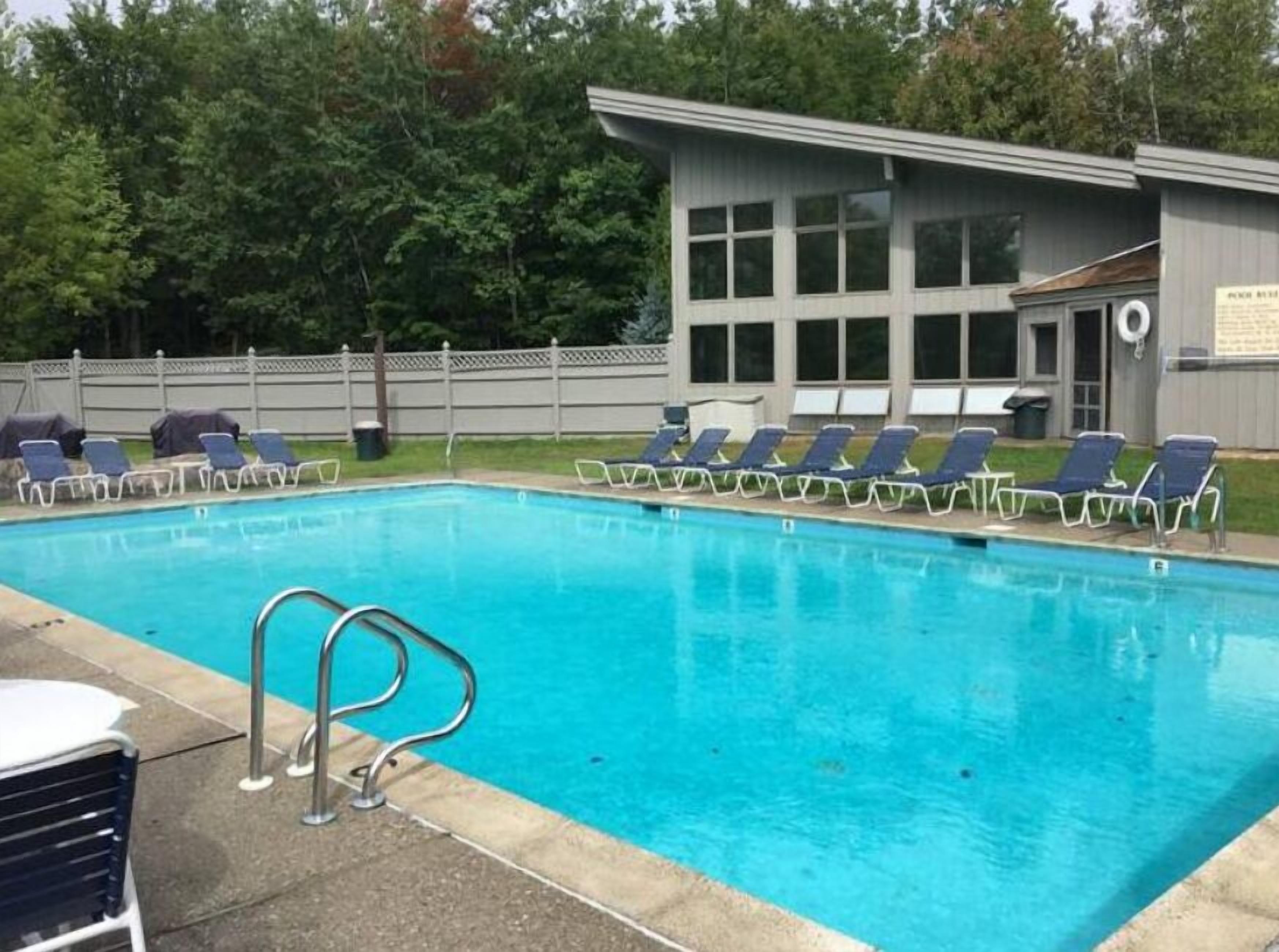 Una piscina rectangular con agua azul cristalina, rodeada de sillas de descanso y un edificio con amplias ventanas al fondo.