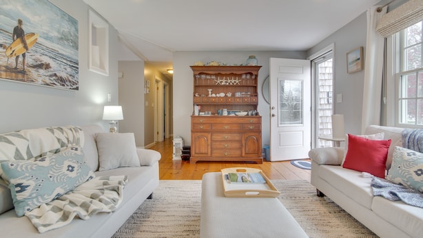 A living room with two white sofas, a wooden cabinet, and a painting of a surfer. - Rail Trail Access: Home 2 Mi to Lighthouse Beach! (Chatham)