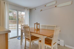 A dining area with a wooden table and chairs, a sliding glass door leading to a balcony, and a wall-mounted paddleboard. - Rail Trail Access: Home 2 Mi to Lighthouse Beach! (Chatham)