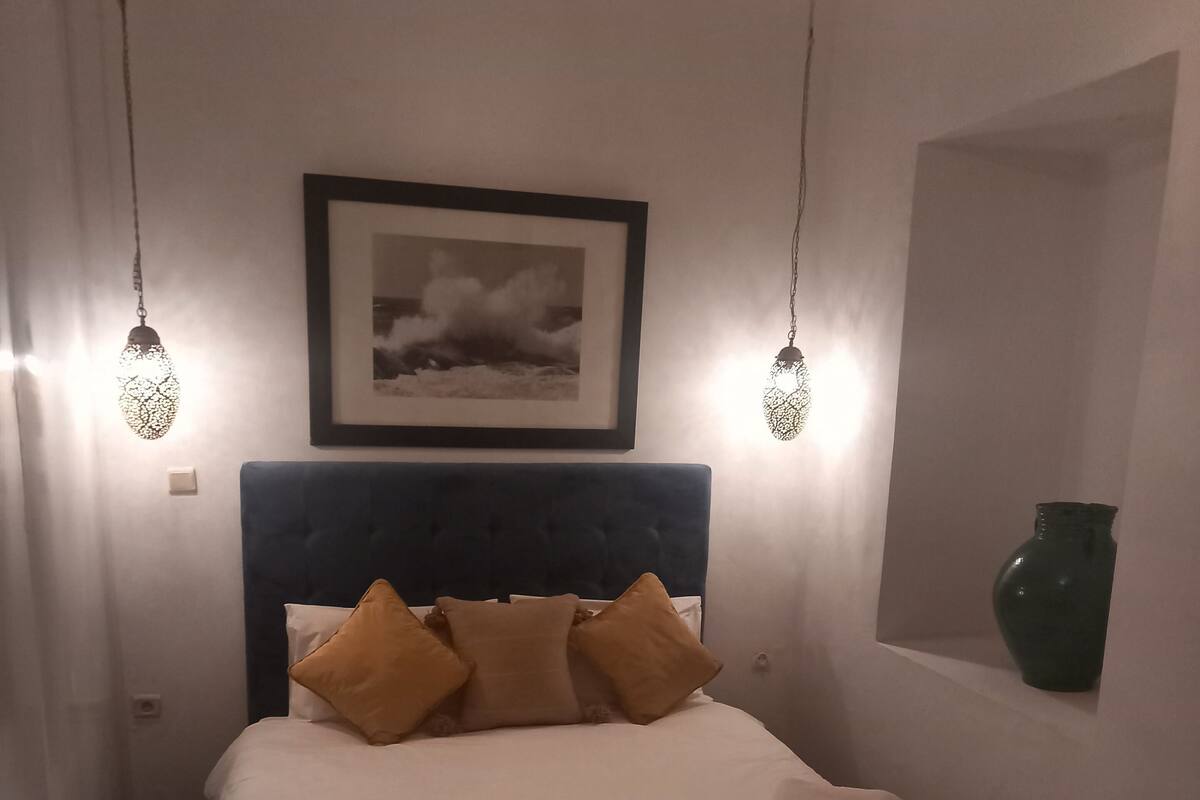 Un lit bien fait, avec une literie blanche et marron, un tableau encadré au-dessus de la tête de lit et deux luminaires suspendus.