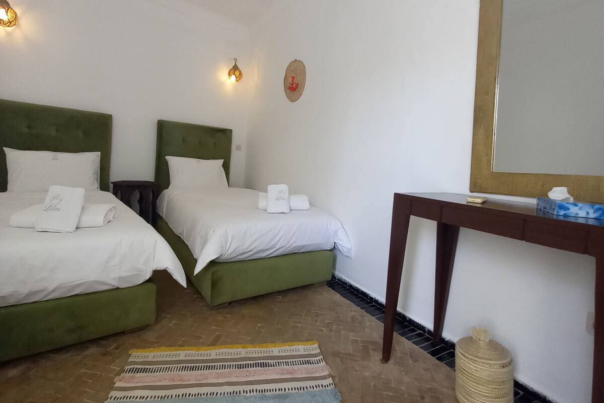Une chambre d’hôtel avec deux lits recouverts d’un tissu vert, du linge de lit blanc, un tapis à motifs rayés, un bureau en bois, un miroir et un appareil de climatisation.