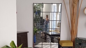 Una habitación con un espejo grande, una planta azul en maceta, un taburete de madera pequeño y un jarrón decorativo con tallos altos.
