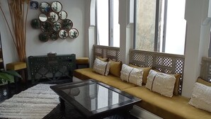 Una sala de estar moderna con un sofá, un reposapiés y una mesa de centro de vidrio. Hay ventanas grandes y un lámpara colgante.
