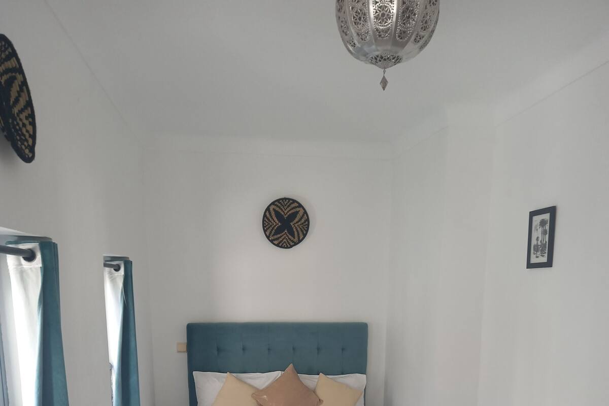 Une chambre à coucher bien rangée, avec un lit, des oreillers, une table de chevet, une horloge murale et une fenêtre avec des rideaux.