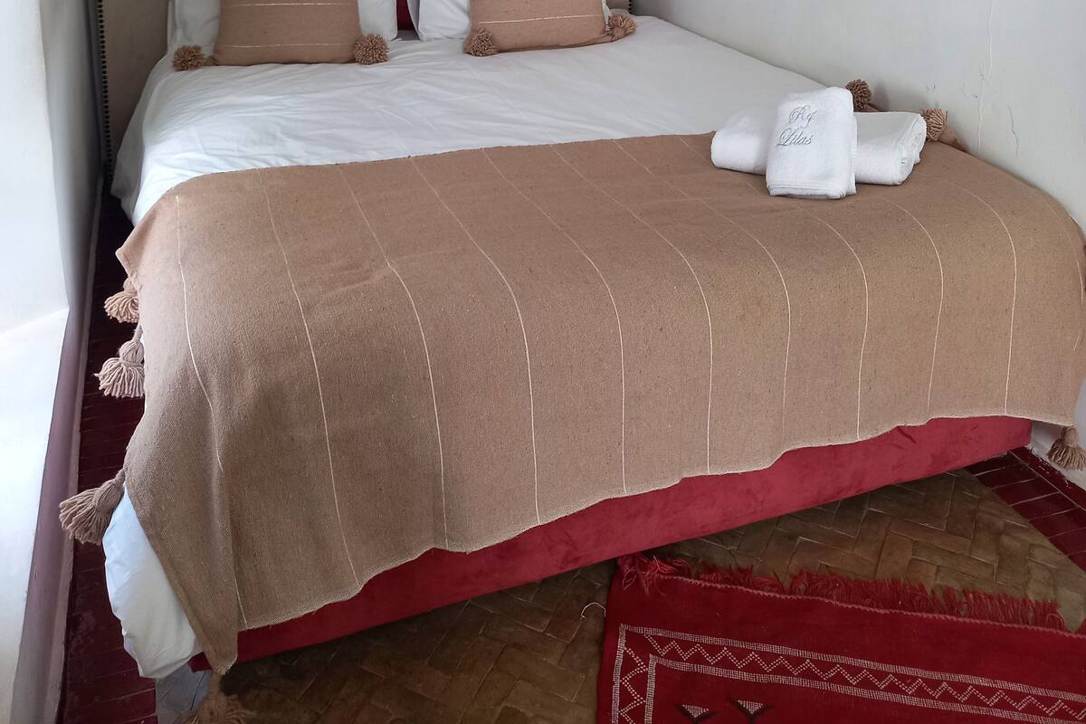Un lit bien fait, avec une tête de lit rouge, une literie blanche et beige, et un plaid marron.