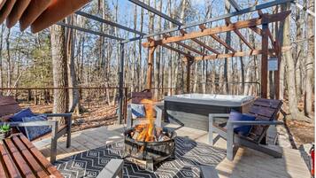 Una terraza con una fogata, asientos y un jacuzzi en un área boscosa.