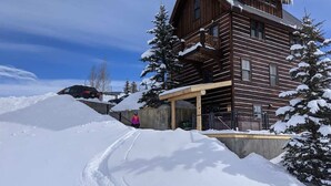 Una cabaña de madera con porche cubierto, árboles nevados y una pista de esquí.