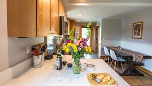 Una cocina moderna con armarios de madera, encimera de mármol, comedor con una mesa larga y sillas, y un jarrón con flores.
