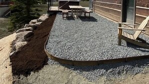 Una cabaña de troncos con un patio de grava, mobiliario exterior y un paisaje de montaña.