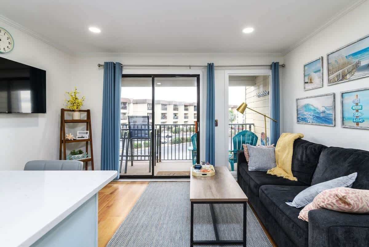 A modern living room with a sofa, a coffee table, and a TV. There is a balcony with blue curtains and a view of buildings.