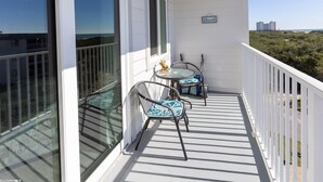 Ein Balkon mit zwei Tischen und Stühlen, großen Fenstern und Blick auf Strand und Gebäude.