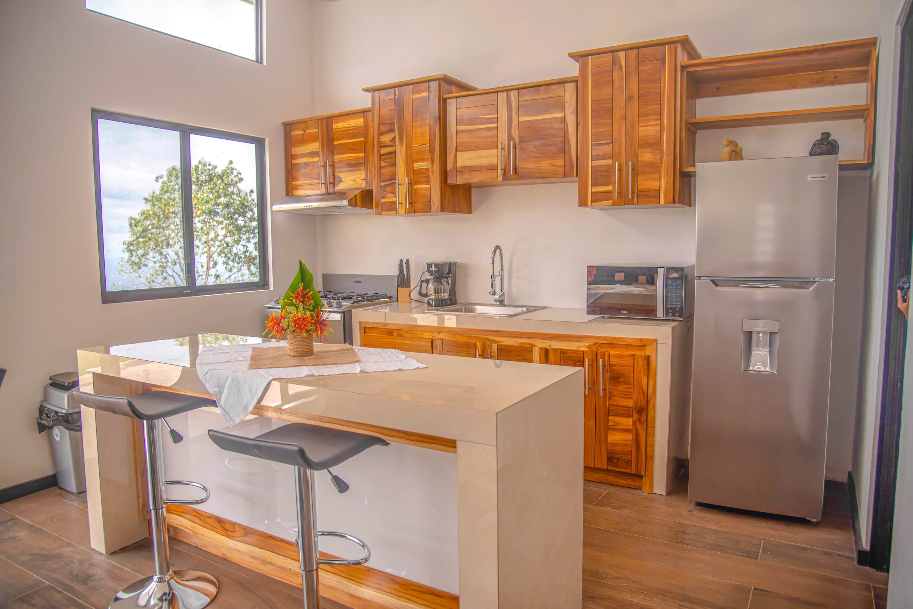 A modern kitchen with wooden cabinets, a stainless steel refrigerator, a built-in island with bar stools, and a window with a view of trees.