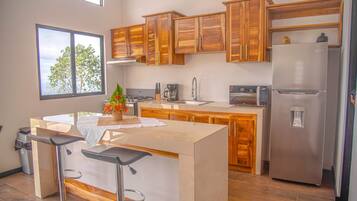 A modern kitchen with wooden cabinets, a stainless steel refrigerator, a built-in island with bar stools, and a window with a view of trees.