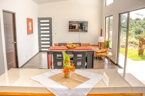 A modern dining area with a wooden table, chairs, and a central island with a fruit bowl. There is a television mounted on the wall and a window providing a view of the outdoors.