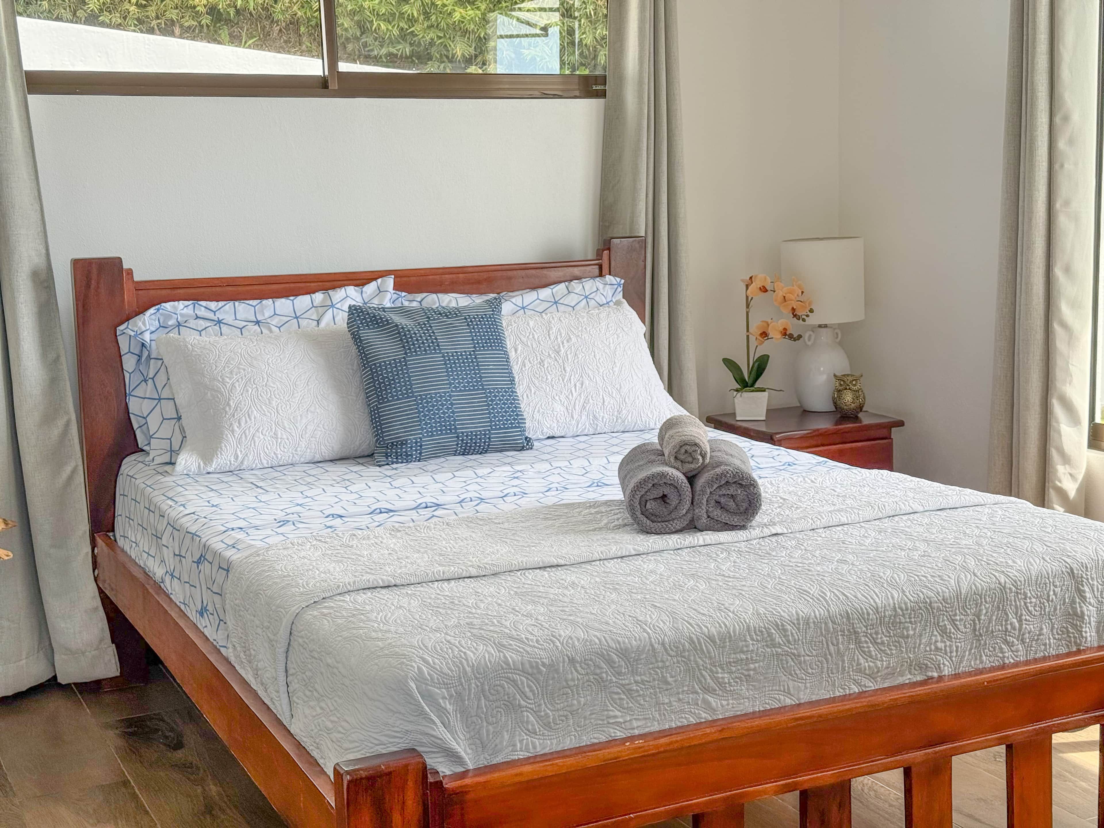 Een netjes opgemaakt bed met behang en kussens, een nachtkastje met een lamp en een klein plantje, en uitzicht op groen buiten.