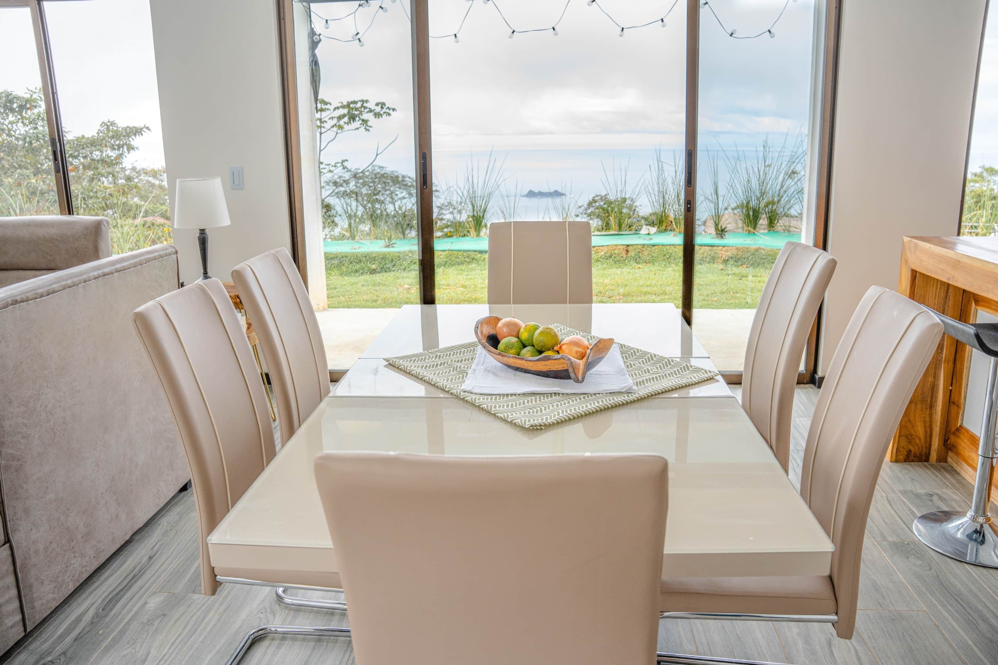A dining area with a table set for four, a bowl of fruit, and a view of the outdoors.