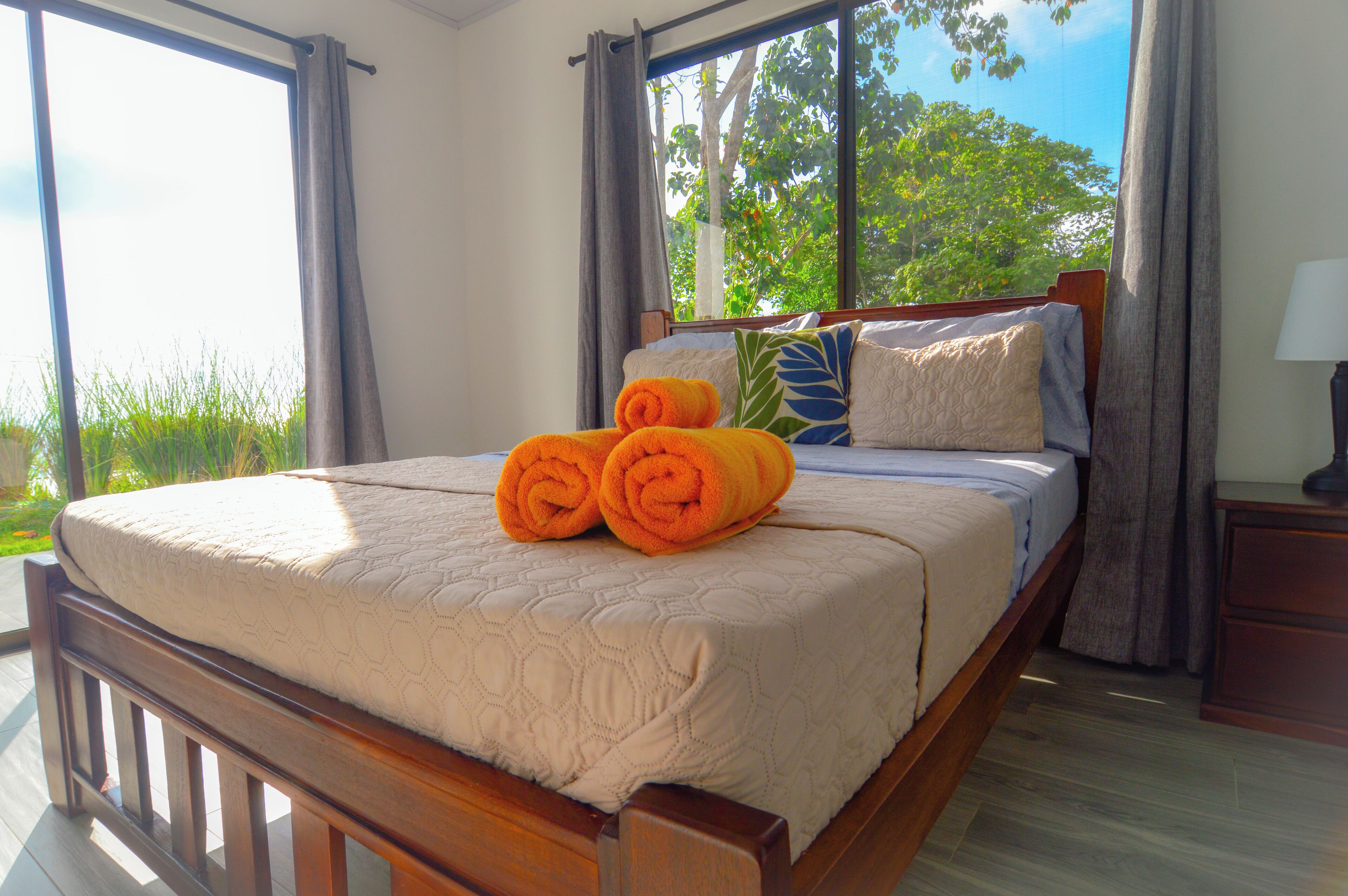 A neatly made bed with a quilt and rolled towels, a wooden headboard, and a view of greenery through large windows.