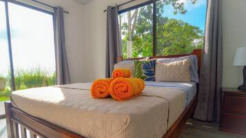 A neatly made bed with a quilt and rolled towels, a wooden headboard, and a view of greenery through large windows.