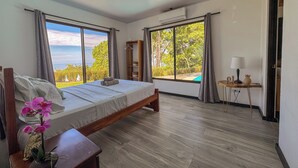 A bedroom with a large bed, a wooden nightstand, a small table with a lamp, and a view of a swimming pool through large windows. - 3bdr House in Uvita LAPA | 180° Ocean View (Bahía Ballena)