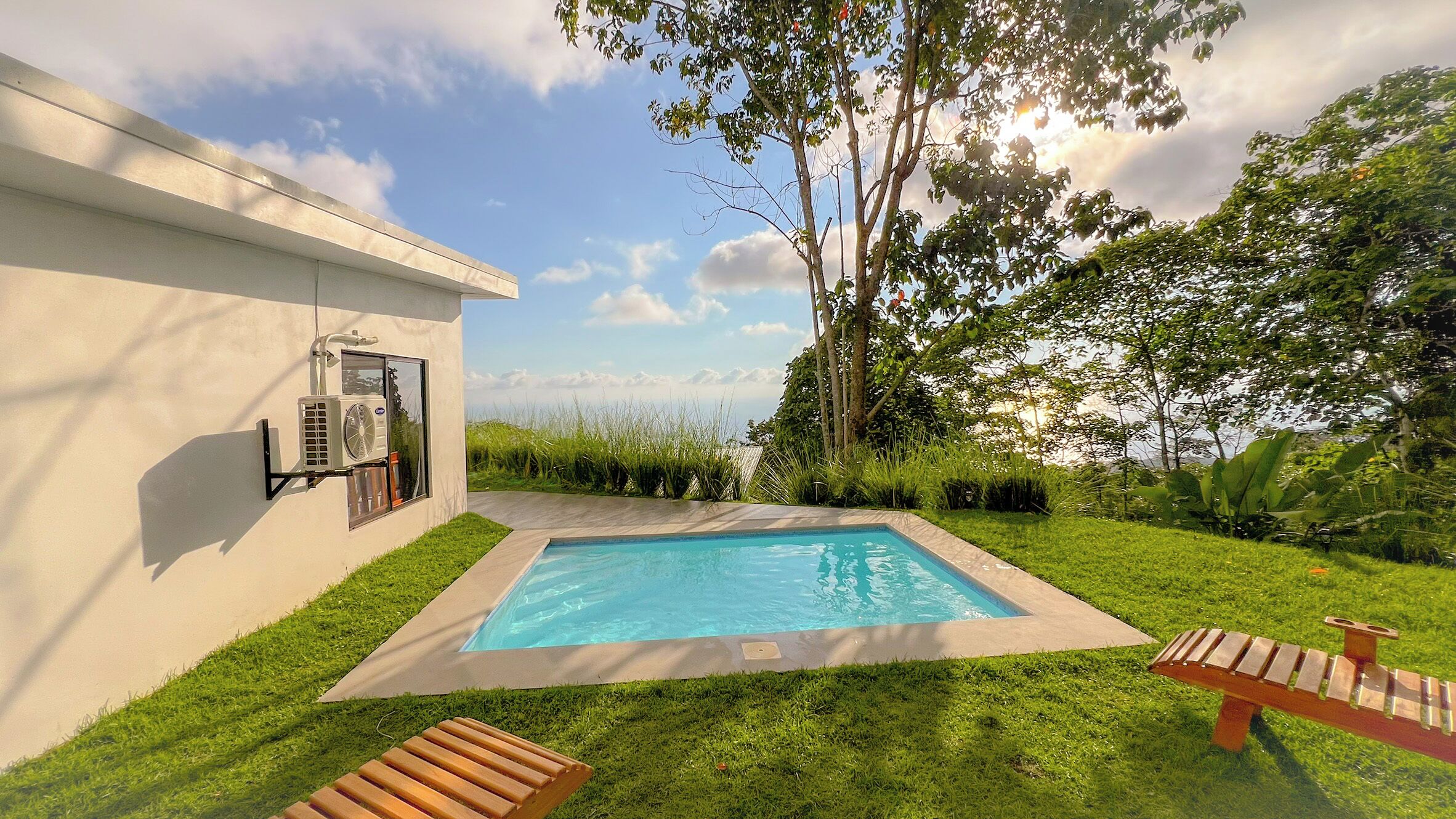 A swimming pool with a wooden deck, surrounded by grass and trees, with a building on one side.
