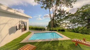 A swimming pool with a wooden deck, surrounded by grass and trees, with a building on one side. - 3bdr House in Uvita LAPA | 180° Ocean View (Bahía Ballena)