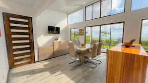 A modern interior with a dining area, a wooden door, and large windows offering a view of greenery. - 3bdr House in Uvita LAPA | 180° Ocean View (Bahía Ballena)
