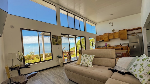 A spacious living room with a large beige sofa, a wooden coffee table, and a view of the ocean through large windows. - 3bdr House in Uvita LAPA | 180° Ocean View (Bahía Ballena)