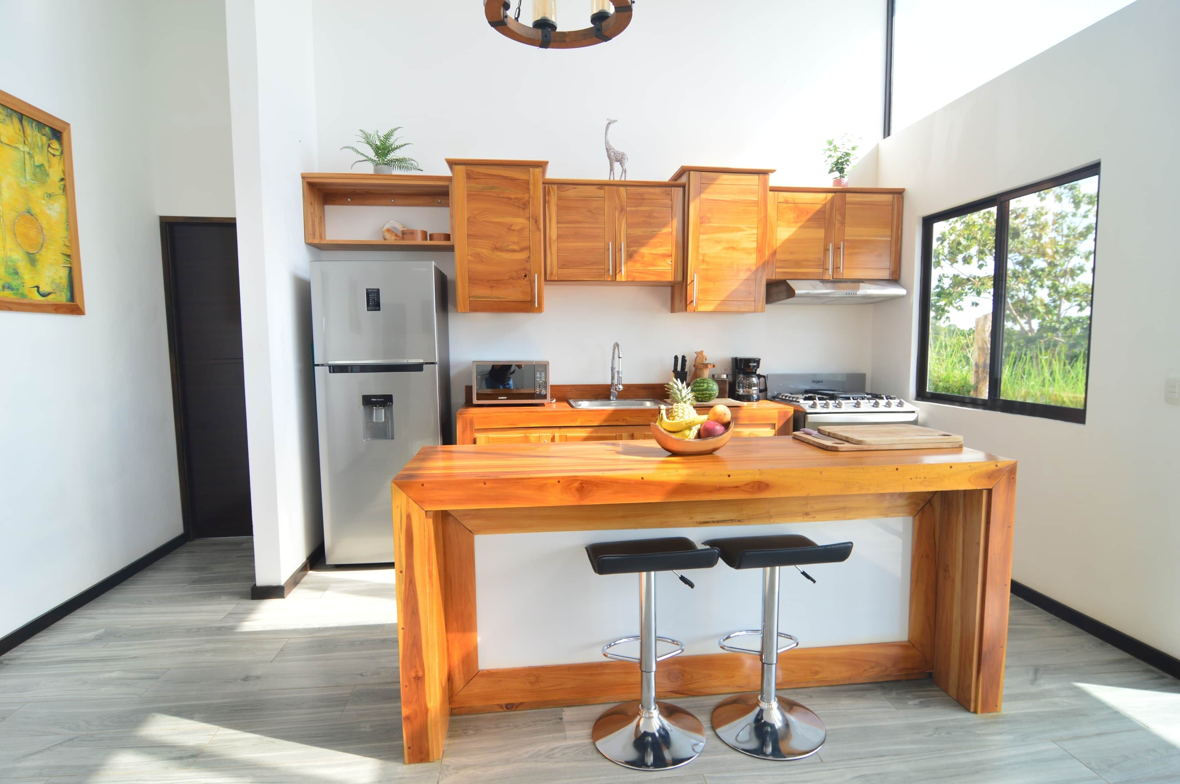 A modern kitchen with wooden cabinets, a central island with stools, a refrigerator, and a window providing a view of greenery.