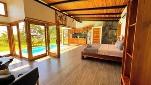 A spacious bedroom with a large bed, wooden flooring, and a view of a swimming pool through glass doors.