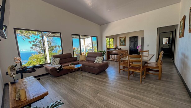 A spacious living area with a large sofa, a dining table with chairs, and a view of the ocean through large windows. - 4bdr House in Uvita QUETZAL | Costa Rica (Uvita)