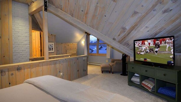 Un dormitorio tipo loft con techo inclinado, vigas de madera, una cama, un televisor y un pequeño rincón de estar.