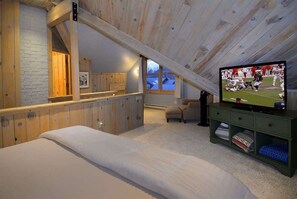A loft bedroom with a sloped ceiling, wooden beams, a bed, a television, and a small seating area.