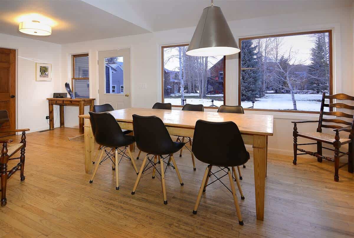 A dining area with a wooden table, black chairs, a pendant light, and large windows with a view of a snowy landscape.
