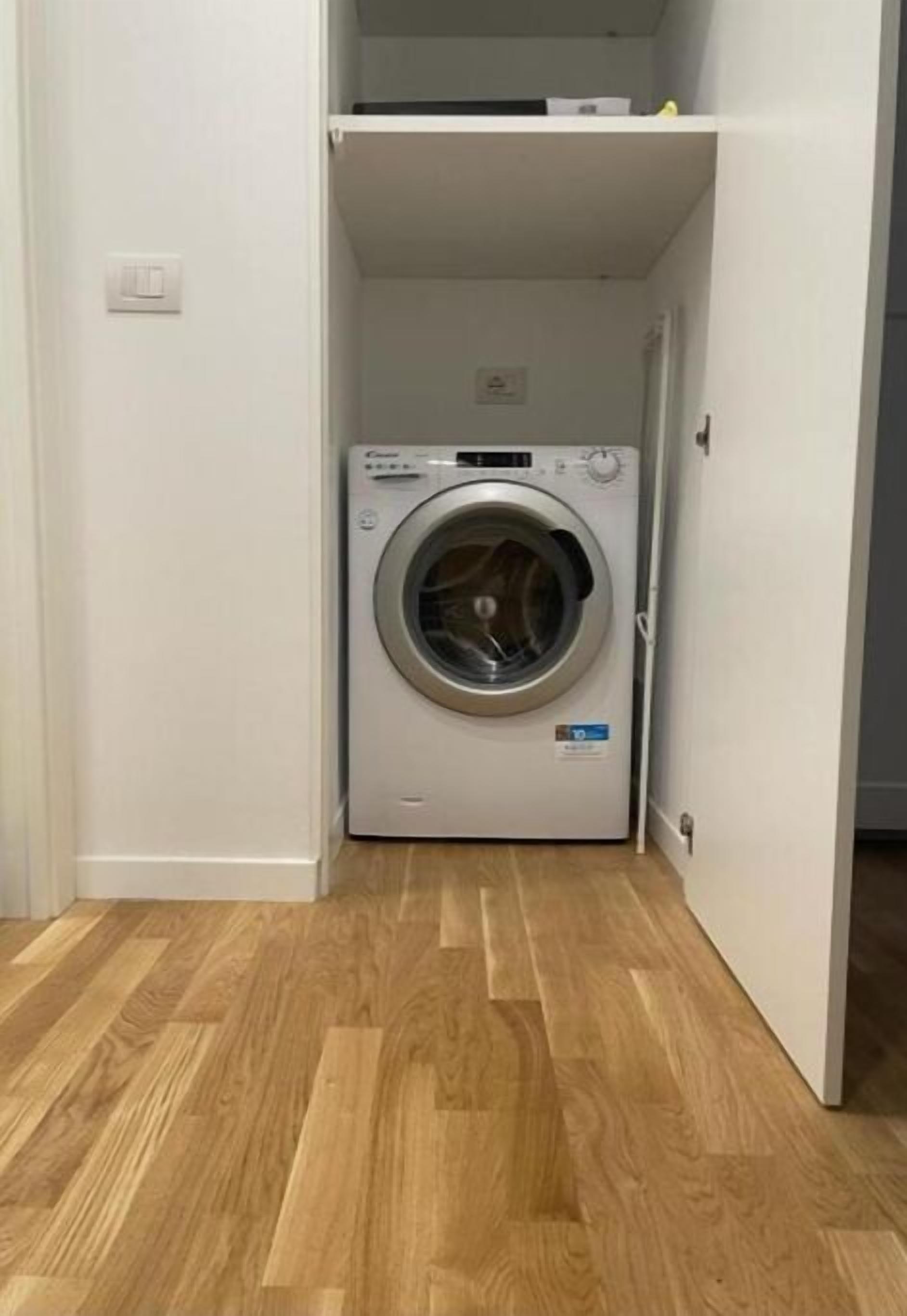 A compact, white washing machine in a small room with wooden flooring and a white door.