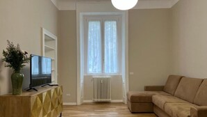 Una sala de estar con un sofá beige, un mueble de madera con diseños geométricos, un televisor sobre un soporte y una ventana con cortinas blancas.