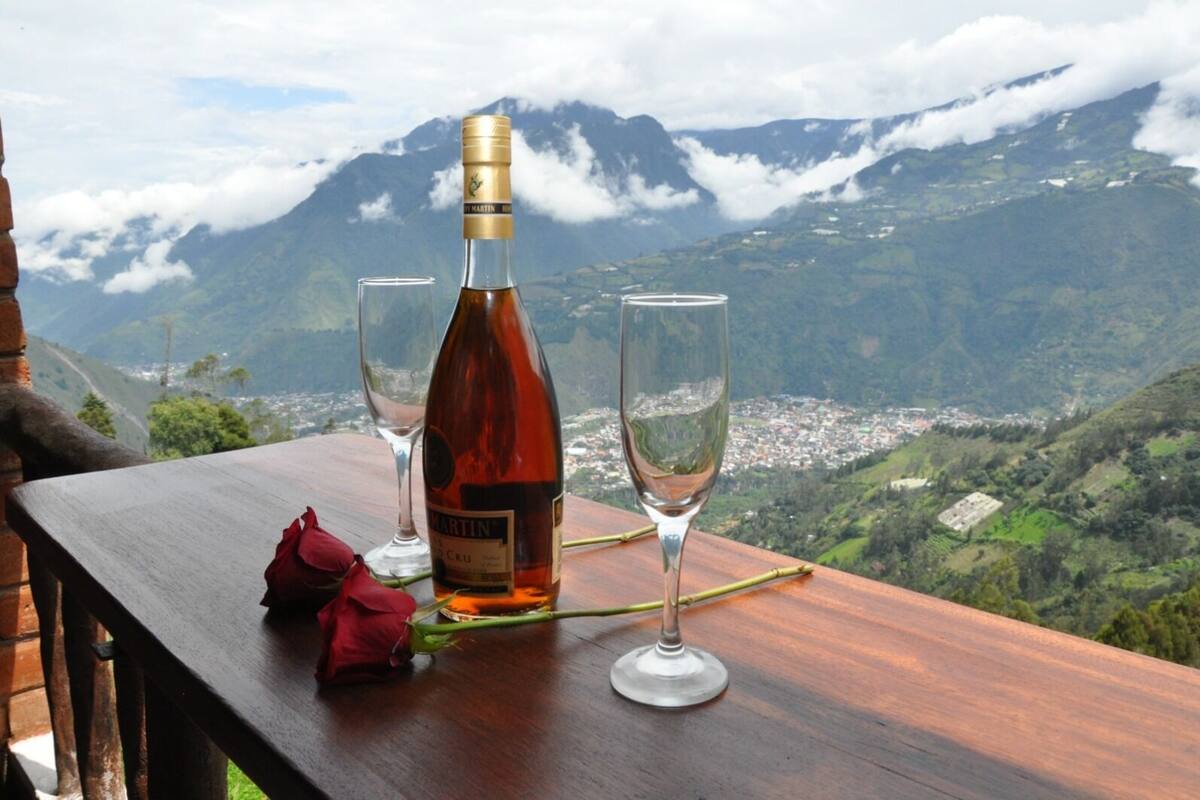 Una botella de vino Martin & Cru sobre una mesa de madera con dos copas, una rosa roja y una vista panorámica de montañas al fondo.