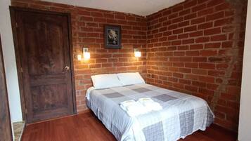 Una habitación con pared de ladrillo, puerta de madera, cama con colcha y dos luces fijadas a la pared.