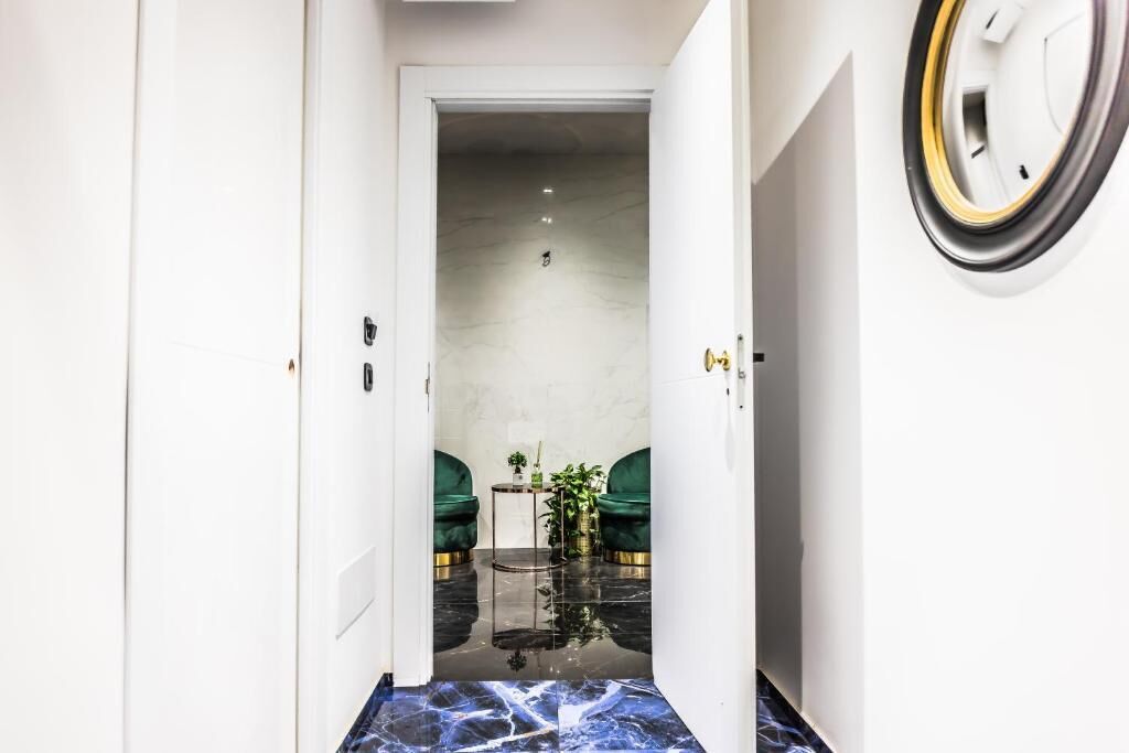 Un couloir au sol en marbre mène à une pièce où se trouvent des fauteuils recouverts d’un tissu vert et une petite table sur laquelle est posée une plante.