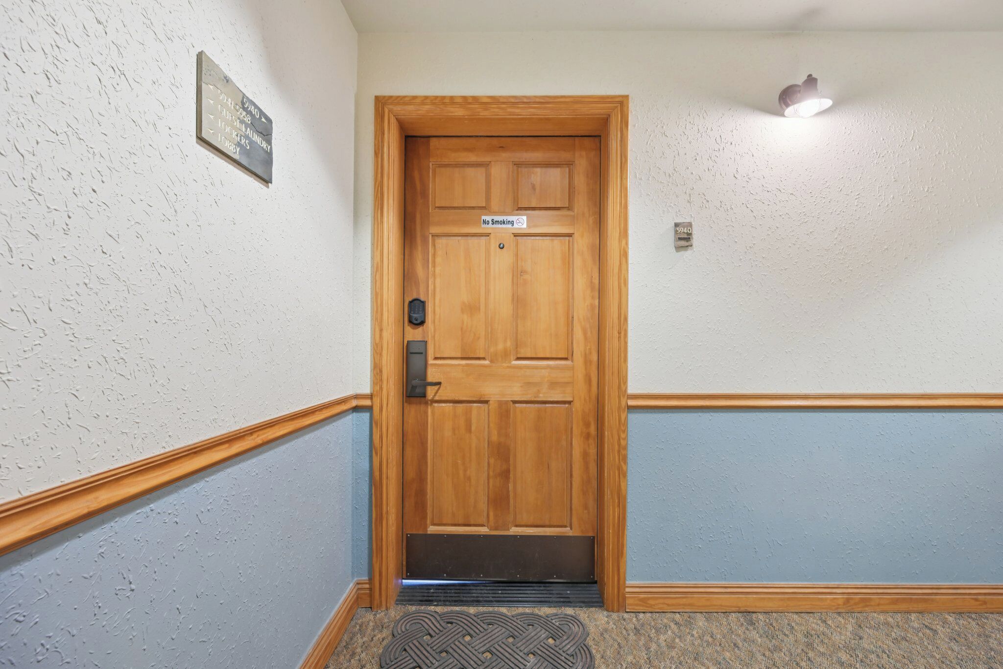 A wooden door with a "No Smoking" sign, a wall-mounted light fixture, and a textured wall with a door mat at the entrance.